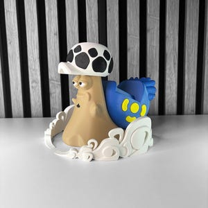 May include: A whimsical, 3D-printed mushroom-shaped figurine. The figurine features a tan body, a white and black spotted cap, and a blue element with yellow spots. It is surrounded by a white, cloud-like base, set against a black and white striped background.