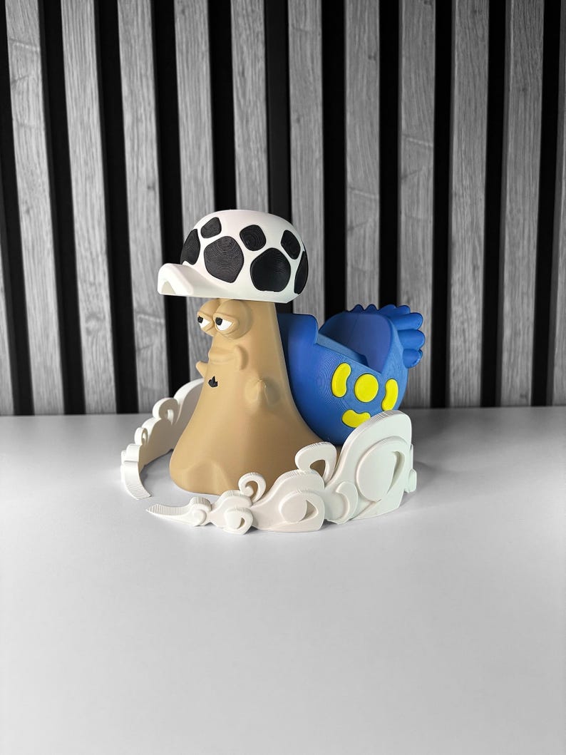 May include: A whimsical, 3D-printed mushroom-shaped figurine. The figurine features a tan body, a white and black spotted cap, and a blue element with yellow spots. It is surrounded by a white, cloud-like base, set against a black and white striped background.