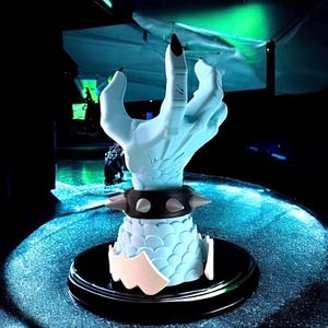 K&ouml;nnte beinhalten: Eine hellblaue, stilisierte Handskulptur mit schwarzen Fingern&auml;geln und einem schwarzen Armband mit Nieten. Die Hand ruht auf einem hellblauen, schuppenartigen Sockel mit schwarzer und wei&szlig;er Zierleiste, auf einem schwarzen kreisf&ouml;rmigen Sockel.