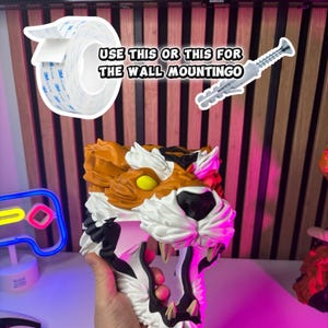 May include: A 3D-printed tiger head with orange, white, and black details, held in a hand. The image includes a roll of double-sided tape and a screw for wall mounting. Text reads "USE THIS OR THIS FOR THE WALL MOUNTINGO."