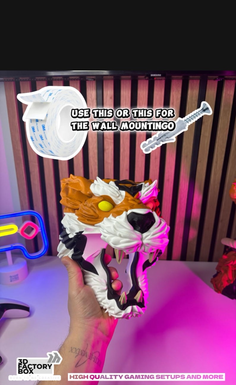 May include: A 3D-printed tiger head with orange, white, and black details, held in a hand. The image includes a roll of double-sided tape and a screw for wall mounting. Text reads "USE THIS OR THIS FOR THE WALL MOUNTINGO."
