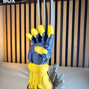 May include: A 3D printed sculpture of a blue and yellow gloved hand with three long, silver-colored claws extending upward. The base features a jagged, gray rock-like formation. The text 3D FACTORY BOX is visible in the upper left corner.
