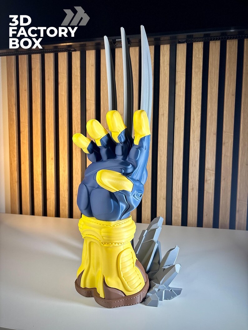 May include: A 3D printed sculpture of a blue and yellow gloved hand with three long, silver-colored claws extending upward. The base features a jagged, gray rock-like formation. The text 3D FACTORY BOX is visible in the upper left corner.