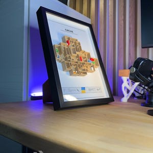May include: A framed 3D map of a video game level, labeled "CACHE". The map is beige with colorful accents and is set against a white background within a black frame. The frame is propped up on a wooden desk.