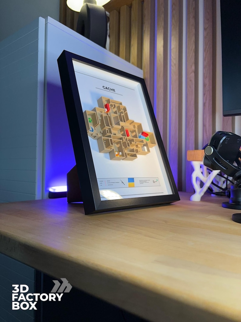 May include: A framed 3D map of a video game level, labeled "CACHE". The map is beige with colorful accents and is set against a white background within a black frame. The frame is propped up on a wooden desk.