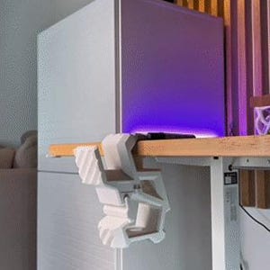 May include: A white, geometric desk clamp is attached to a wooden desk. The clamp is designed to hold items. A white cabinet with horizontal lines is in the background. A lamp with a black shade is above the cabinet.