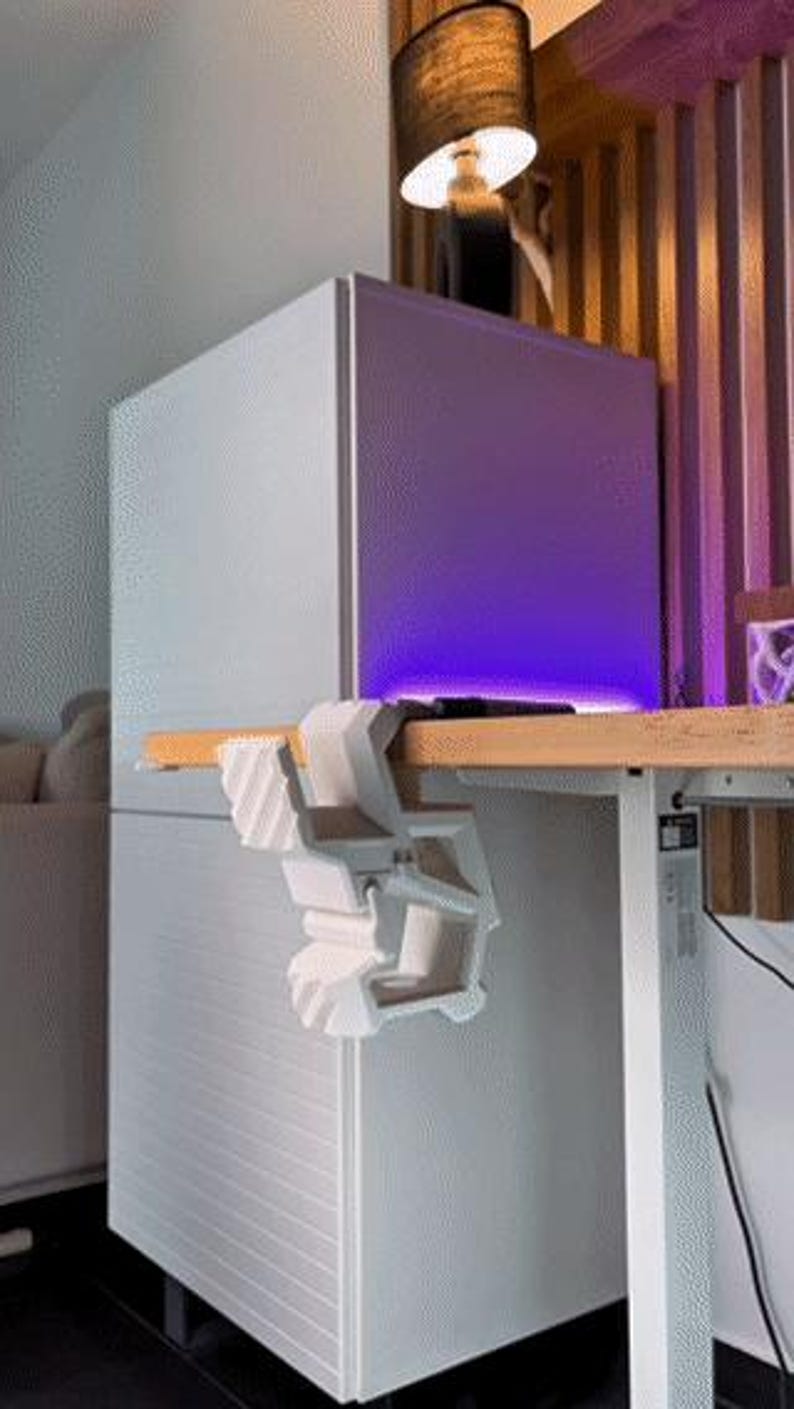 May include: A white, geometric desk clamp is attached to a wooden desk. The clamp is designed to hold items. A white cabinet with horizontal lines is in the background. A lamp with a black shade is above the cabinet.