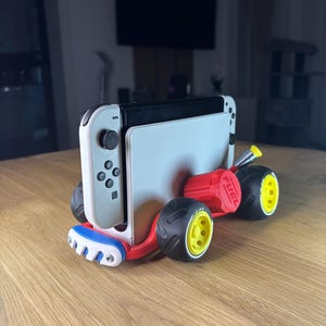 K&ouml;nnte beinhalten: Eine wei&szlig;-schwarze Nintendo Switch-Konsole, die in einen roten, blauen und schwarzen Kart eingebaut ist. Der Kart hat schwarze Reifen mit gelben Felgen und das Wort "FUEL" auf dem roten Motor aufgedruckt.