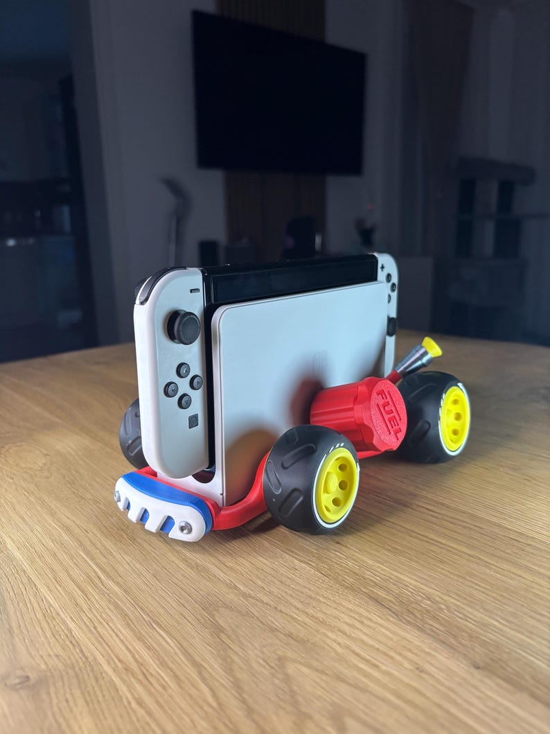 K&ouml;nnte beinhalten: Eine wei&szlig;-schwarze Nintendo Switch-Konsole, die in einen roten, blauen und schwarzen Kart eingebaut ist. Der Kart hat schwarze Reifen mit gelben Felgen und das Wort "FUEL" auf dem roten Motor aufgedruckt.