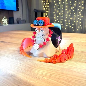 Peut inclure: Une figurine imprim&eacute;e en 3D d'un personnage de dessin anim&eacute; avec un chapeau rouge, des lunettes bleues et un collier de perles rouges. La figurine est debout sur une base avec des flammes orange.