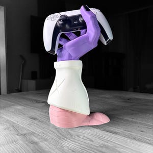 Op de afbeelding: Een sculptuur van een paarse hand die een witte en zwarte gamecontroller vasthoudt. De hand is bevestigd aan een witte en roze basis, waardoor een uniek decoratief stuk voor gamers ontstaat.