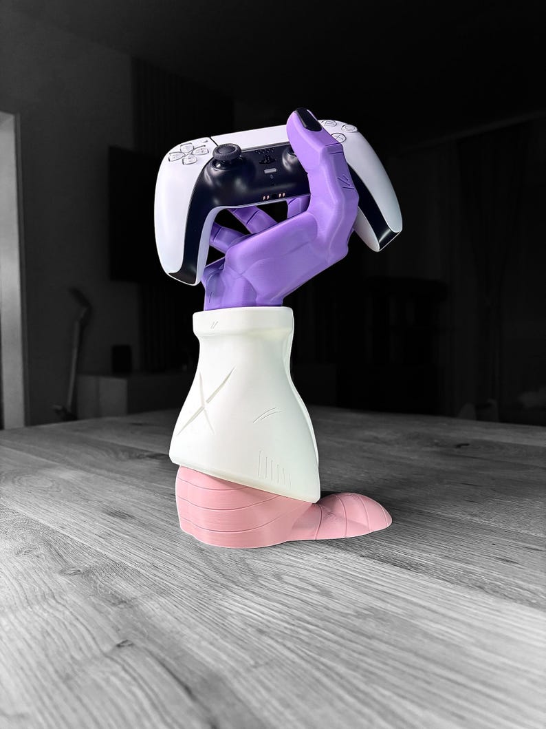 Op de afbeelding: Een sculptuur van een paarse hand die een witte en zwarte gamecontroller vasthoudt. De hand is bevestigd aan een witte en roze basis, waardoor een uniek decoratief stuk voor gamers ontstaat.