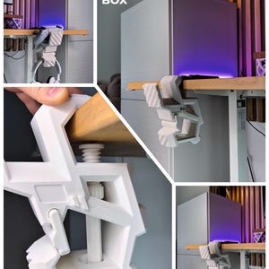 May include: A white 3D-printed headphone holder clamped to a wooden desk. The holder is supporting a pair of white headphones. The text "3D FACTORY BOX" is visible in the image. A computer tower with purple lighting is also visible.
