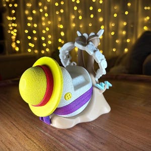 May include: A novelty figurine of a snail wearing a yellow hat with a red band and purple detailing. The snail's shell is white with a button. The figurine is tan and has a cartoon design.