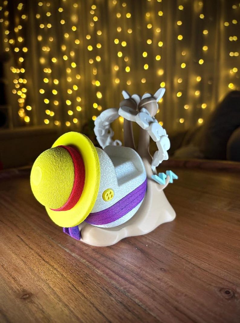 May include: A novelty figurine of a snail wearing a yellow hat with a red band and purple detailing. The snail's shell is white with a button. The figurine is tan and has a cartoon design.