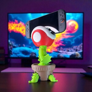 Könnte beinhalten: Ein neuartiger Telefon- oder Spielekonsolenhalter in Form einer rot-weißen Piranha-Pflanze aus der Super Mario-Videospielserie. Der Halter befindet sich in einem braunen Topf mit grünen Akzenten und hält eine schwarze Nintendo Switch-Konsole.