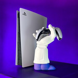Puede incluir: Una consola PlayStation 5 blanca con una mano blanca sosteniendo un mando PlayStation 5 blanco. La mano lleva un guante azul y est&aacute; de pie sobre una superficie morada.