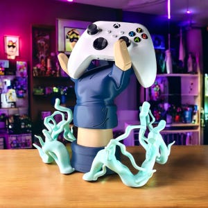 May include: A white Xbox controller held by a blue and tan figurine. The figurine has a blue sleeve and tan arm. The base of the figurine has a light blue, swirling design. The controller has colorful buttons.