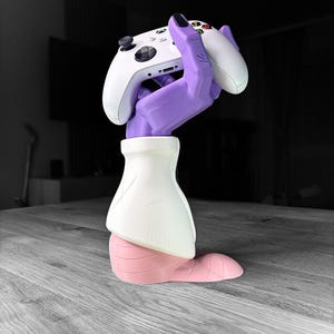 Op de afbeelding: Een witte Xbox-controller die wordt vastgehouden door een paarse handvormige standaard. De standaard heeft een witte mouw en een roze basis. De controller heeft zwarte knoppen en een D-pad. De achtergrond is wazig.