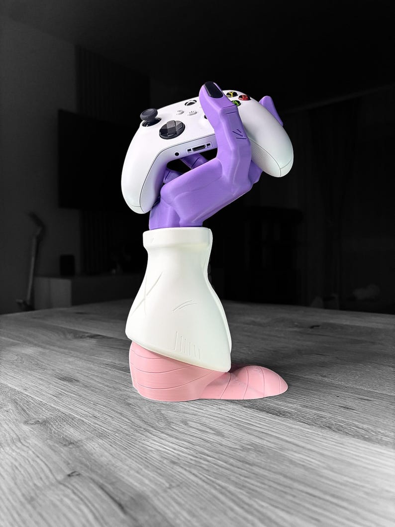 Op de afbeelding: Een witte Xbox-controller die wordt vastgehouden door een paarse handvormige standaard. De standaard heeft een witte mouw en een roze basis. De controller heeft zwarte knoppen en een D-pad. De achtergrond is wazig.