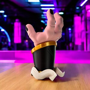 May include: A sculpture of a hand with black fingernails extending from a black and gold cuff. The cuff sits atop a black cylindrical base with a white, curved, decorative element at the bottom. The background is blurred with purple and blue lights.