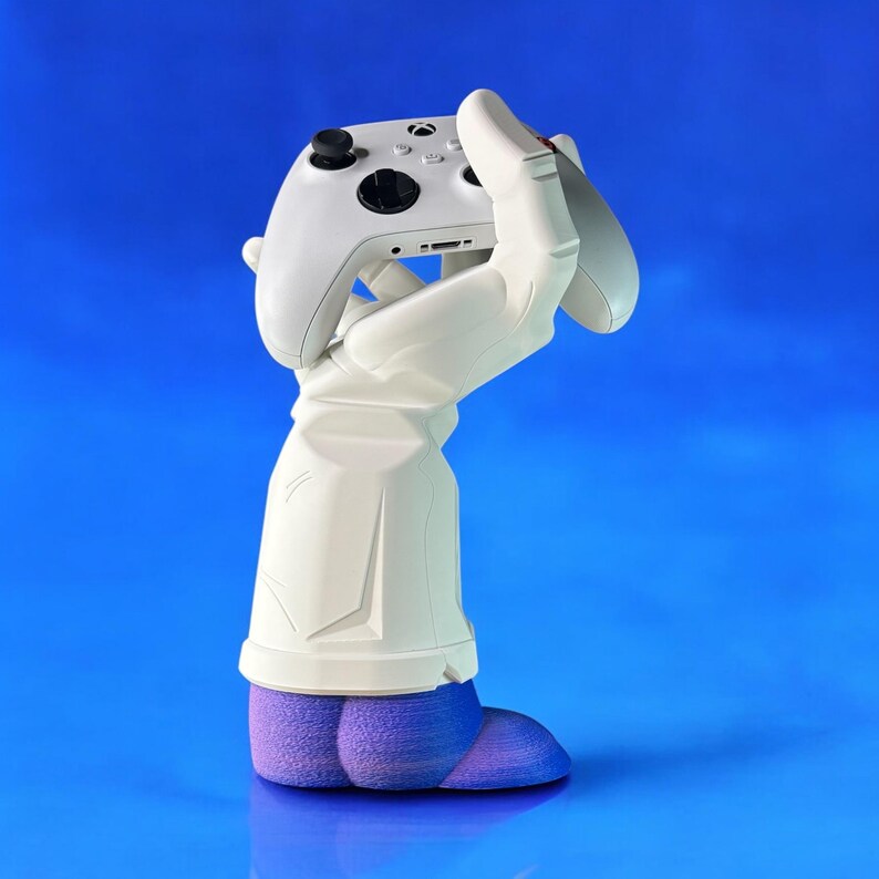Puede incluir: Una mano blanca impresa en 3D sosteniendo un mando de videojuego blanco. La mano lleva un zapato morado y azul.