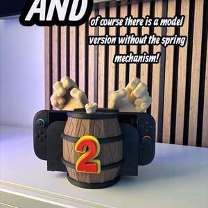 May include: A black Nintendo Switch controller holder shaped like a wooden barrel with a large red number 2. The text "AND" and "of course there is a model version without the spring mechanism!" are also visible.
