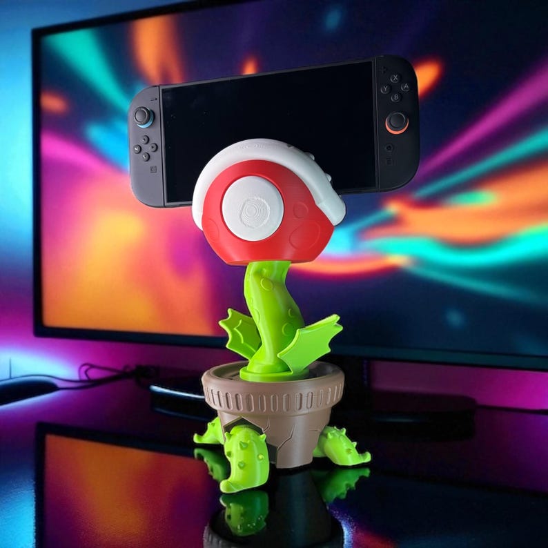 May include: A novelty Nintendo Switch stand shaped like a Piranha Plant from the Super Mario video game series. The stand is red, white, and green, with a brown pot base. The Switch console is held in the plant's mouth.