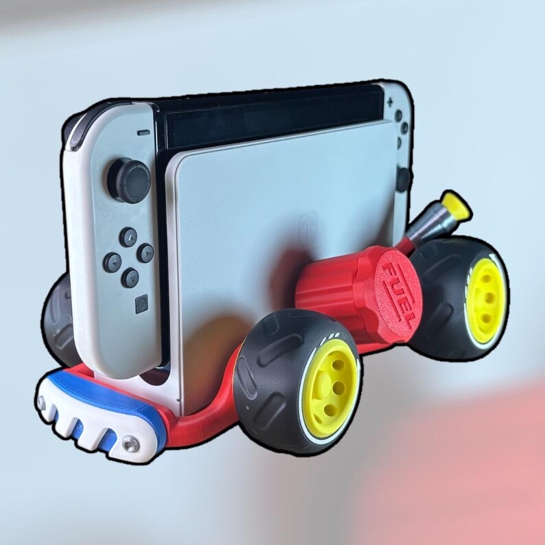 K&ouml;nnte beinhalten: Eine wei&szlig;e und schwarze Nintendo Switch-Konsole, die an einem rot-schwarz-gelben Zubeh&ouml;r in Form eines Karts befestigt ist. Das Zubeh&ouml;r hat R&auml;der und einen roten Tankdeckel mit dem Aufdruck "FUEL". Das Kart hat eine blau-wei&szlig;-rote Frontsto&szlig;stange.