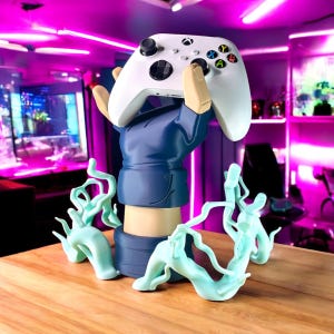 May include: A white Xbox controller is held aloft by a blue-sleeved arm statue. The statue has a tan arm and hand, with light blue decorative accents. The controller has colorful buttons and black joysticks. The statue sits on a wooden surface.