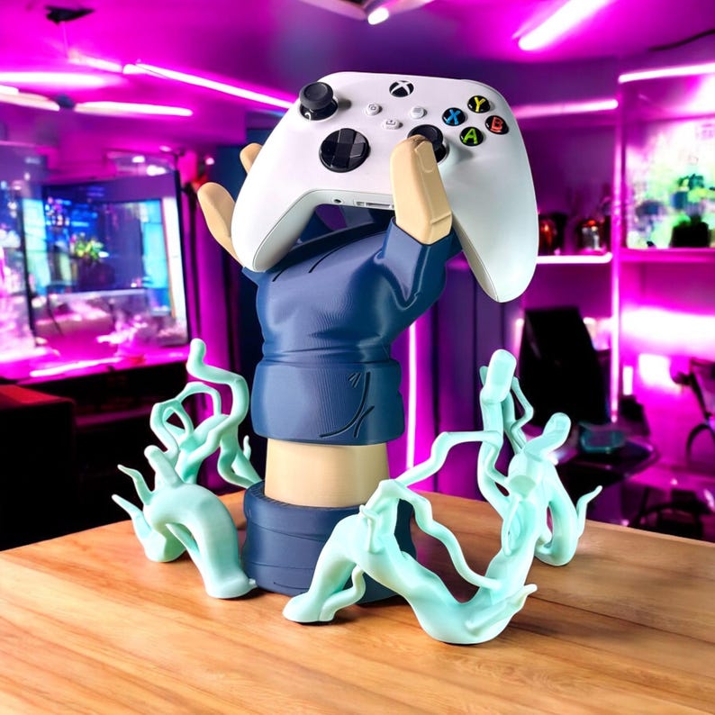 May include: A white Xbox controller is held aloft by a blue-sleeved arm statue. The statue has a tan arm and hand, with light blue decorative accents. The controller has colorful buttons and black joysticks. The statue sits on a wooden surface.