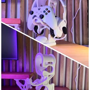 Peut inclure: Support blanc imprim&eacute; en 3D pour casque et manette de jeu. Le support maintient un casque et une manette blancs. Le texte "3D FACTORY BOX" est dans le coin sup&eacute;rieur gauche. Le support est sur une surface en bois.