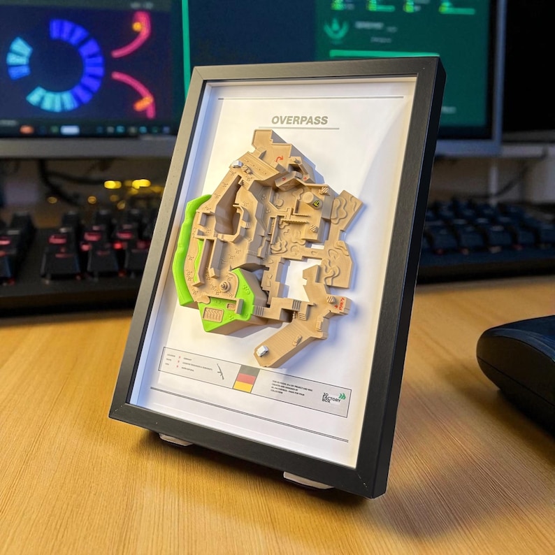 May include: A framed 3D map of the Overpass game level, featuring tan and green terrain details. The black frame holds the map, with the word "OVERPASS" at the top. The map is set on a wooden desk.
