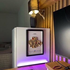 May include: A black table lamp with a cylindrical base and a black lampshade. The lamp sits atop a white cabinet, with a framed artwork and the text "CACHE" visible. The image also shows a wooden shelf and a desk.
