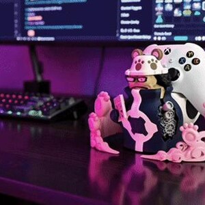 Puede incluir: Un soporte para mando de videojuegos con un dise&ntilde;o de personaje que sostiene un mando blanco. El soporte es principalmente rosa y azul marino, con un mando blanco. Un monitor de ordenador est&aacute; en el fondo. El escritorio es de madera oscura.