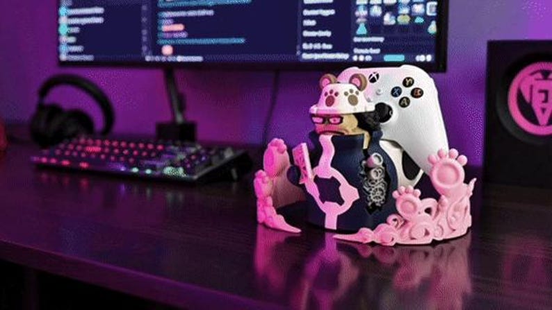 Puede incluir: Un soporte para mando de videojuegos con un dise&ntilde;o de personaje que sostiene un mando blanco. El soporte es principalmente rosa y azul marino, con un mando blanco. Un monitor de ordenador est&aacute; en el fondo. El escritorio es de madera oscura.