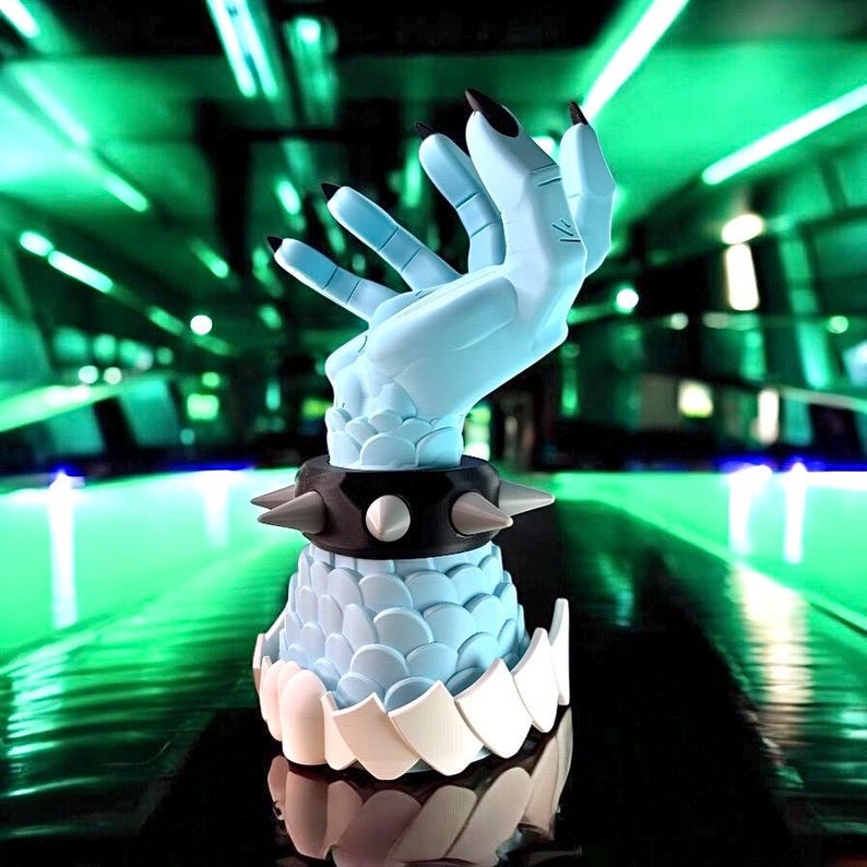 K&ouml;nnte beinhalten: Eine hellblaue Skulptur einer Hand mit langen schwarzen Fingern&auml;geln. Die Hand ist erhoben, und das Handgelenk ist mit einem schwarzen, mit Nieten besetzten Kragen verziert. Die Basis der Skulptur hat ein schuppenartiges Muster und einen wei&szlig;en, zahnartigen Besatz.