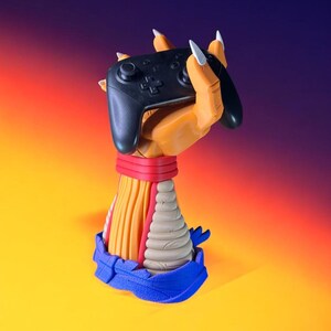 Puede incluir: Un mando de videojuegos negro sostenido por una mano naranja esculpida con garras blancas. La mano forma parte de una escultura colorida con una banda roja, detalles amarillos y beige y una base azul. El fondo hace una transici&oacute;n de amarillo a naranja y morado.
