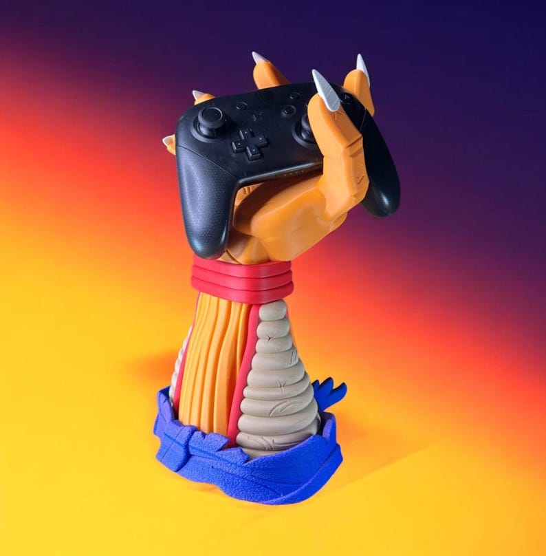Puede incluir: Un mando de videojuegos negro sostenido por una mano naranja esculpida con garras blancas. La mano forma parte de una escultura colorida con una banda roja, detalles amarillos y beige y una base azul. El fondo hace una transici&oacute;n de amarillo a naranja y morado.