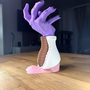 Op de afbeelding: Een sculptuur van een hand met een paarse handpalm en zwarte nagels. De sculptuur heeft een witte en bruine basis en een roze voetachtige verlenging. Het kunstwerk staat op een houten tafel.