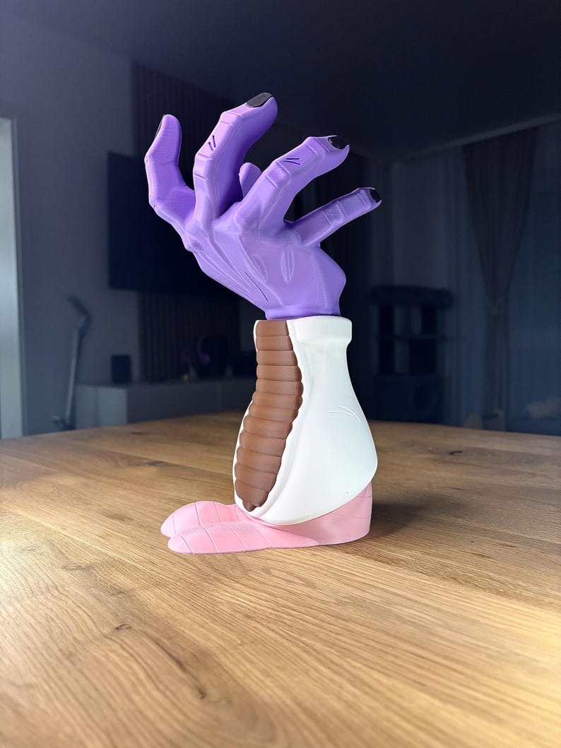 Op de afbeelding: Een sculptuur van een hand met een paarse handpalm en zwarte nagels. De sculptuur heeft een witte en bruine basis en een roze voetachtige verlenging. Het kunstwerk staat op een houten tafel.