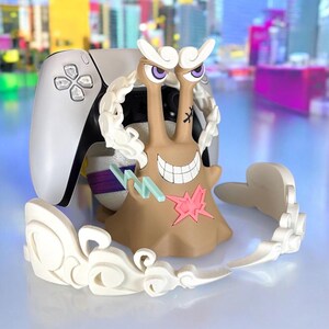 May include: A tan and white figurine of a snail-like character with a wide grin, purple eyes, and white cloud-like accents. The figurine is next to a white and black game controller, with a city backdrop.
