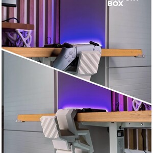 May include: A white and gray gaming controller is held by a white 3D-printed holder attached to a wooden desk. The image includes the text "3D FACTORY BOX". The background features a purple glow and wooden paneling.