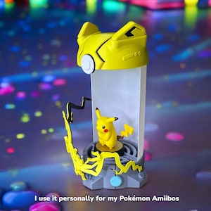 May include: A yellow and gray Pokemon figure display stand with a clear plastic case. The stand features a Pikachu figure inside the case. The stand has a lightning bolt design and a blue button.