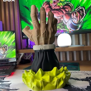 Op de afbeelding: Een 3D-geprinte sculptuur van een hand met een zwarte basis en een geel, stekelig, energie-achtig ontwerp. De hand is beige en lijkt een gebaar te maken. De tekst "3D FACTORY BOX" is zichtbaar in de linkerbovenhoek.