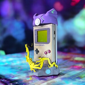 K&ouml;nnte beinhalten: Ein violetter und gr&uuml;ner 3D-gedruckter St&auml;nder mit einem gelben Design h&auml;lt einen Vintage Game Boy mit einem violetten und gr&uuml;nen 3D-gedruckten Geh&auml;use. Der Game Boy steht auf einer bunten Oberfl&auml;che.