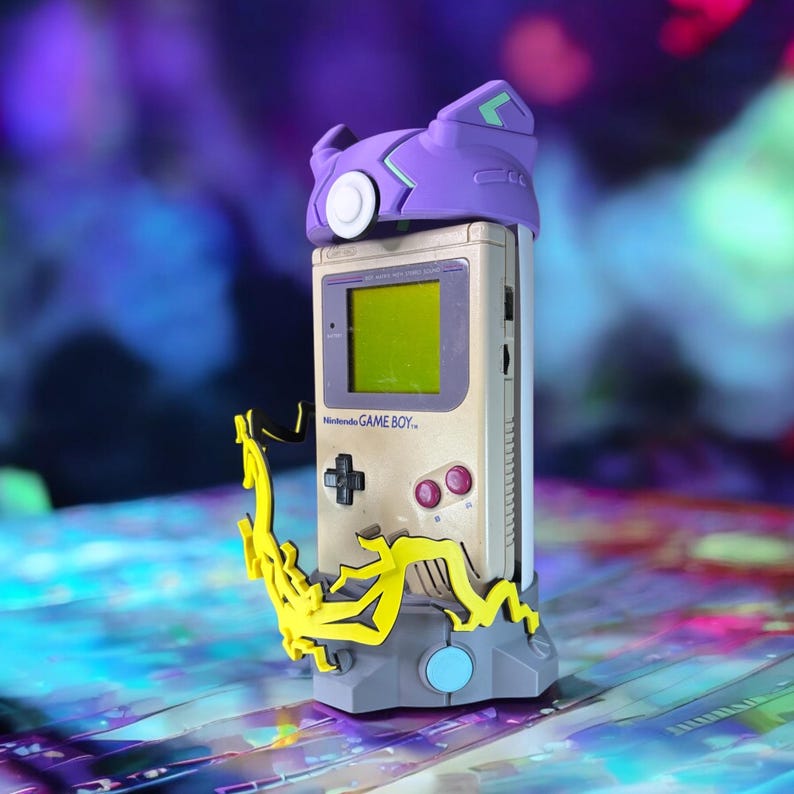 K&ouml;nnte beinhalten: Ein violetter und gr&uuml;ner 3D-gedruckter St&auml;nder mit einem gelben Design h&auml;lt einen Vintage Game Boy mit einem violetten und gr&uuml;nen 3D-gedruckten Geh&auml;use. Der Game Boy steht auf einer bunten Oberfl&auml;che.