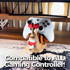 May include: A white Xbox controller rests on a whimsical, cartoonish snail-shaped stand. The snail has a smiling face, heart-shaped eyes, and orange and black stripes. The text "Compatible to ALL Gaming Controller!" is displayed.
