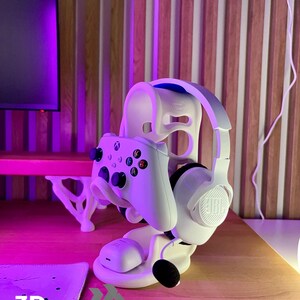 Peut inclure: Support blanc pour casque et manette de jeu. Le support maintient une manette Xbox blanche et un casque JBL blanc. La base porte le texte "3D FACTORY BOX". Le support est sur une surface en bois.