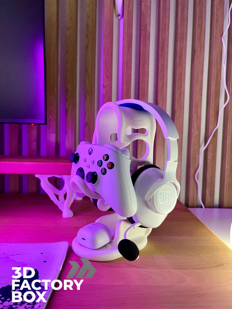 Peut inclure: Support blanc pour casque et manette de jeu. Le support maintient une manette Xbox blanche et un casque JBL blanc. La base porte le texte "3D FACTORY BOX". Le support est sur une surface en bois.